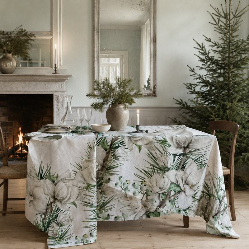 luxury-linen-tablecloth-magnolia-bianca Tovaglia natalizia in lino Magnolia di La Bottega di Casa su tavola apparecchiata con caraffa, calici e vaso di rami di pino — magnolie bianche e bacche verdi lungo i lati, davanti a camino acceso, specchiera antica e albero di Natale