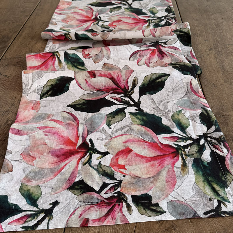 runner-da-tavola-magnolia-rosa3-La-Bottega-di-Casa Runner in lino Magnolia Rosa in primo piano su tavolo in legno — grandi fiori di magnolia in rosa cipria e cardinale con fogliame verde scuro in acquerello